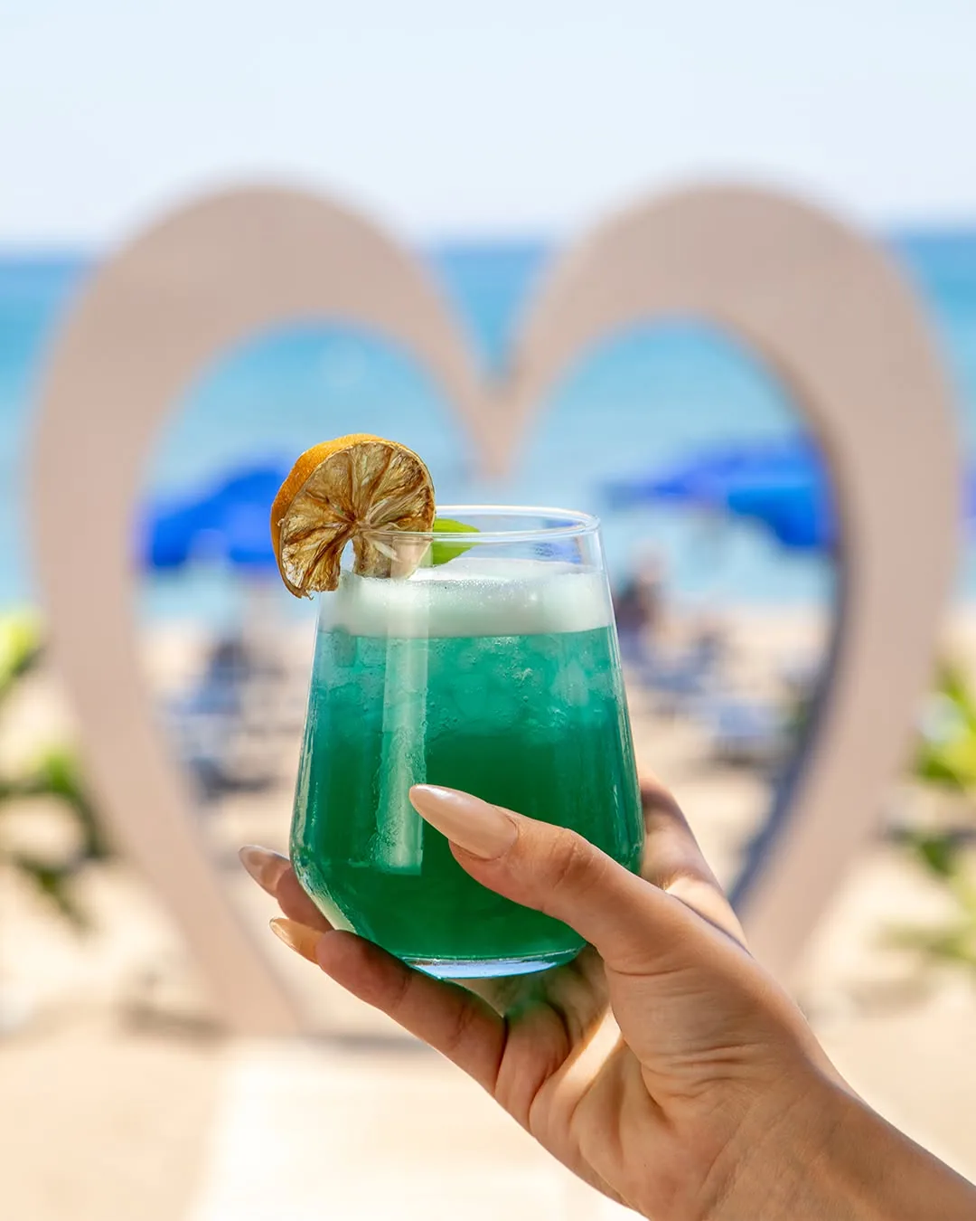 Χέρι κρατά πράσινο cocktail μπροστά από ξύλινο διακοσμητικό σε σχήμα καρδιάς και θέα στην παραλία
